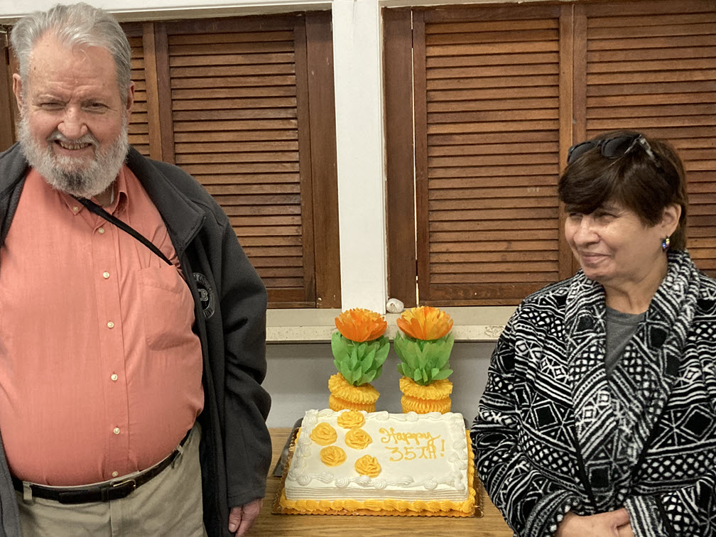 Roger Petersen and Lupe Medrano stand on either side of a large sheet cake. The cake is a white cake with lemon filling, whipped cream frosting and 5 yellow roses. In yellow it says 'Happy 35th!'.