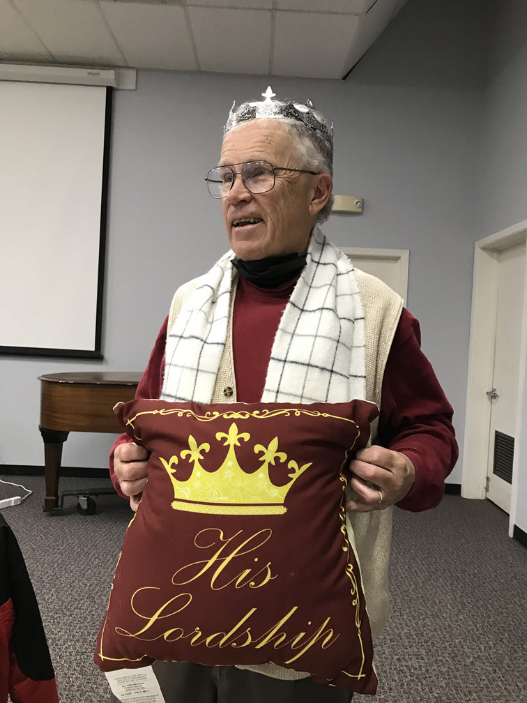 Victor Clifford is shown wearing a silver crown and holding a dark purple pillow that has a crown and the words 'His Lordship' on it.